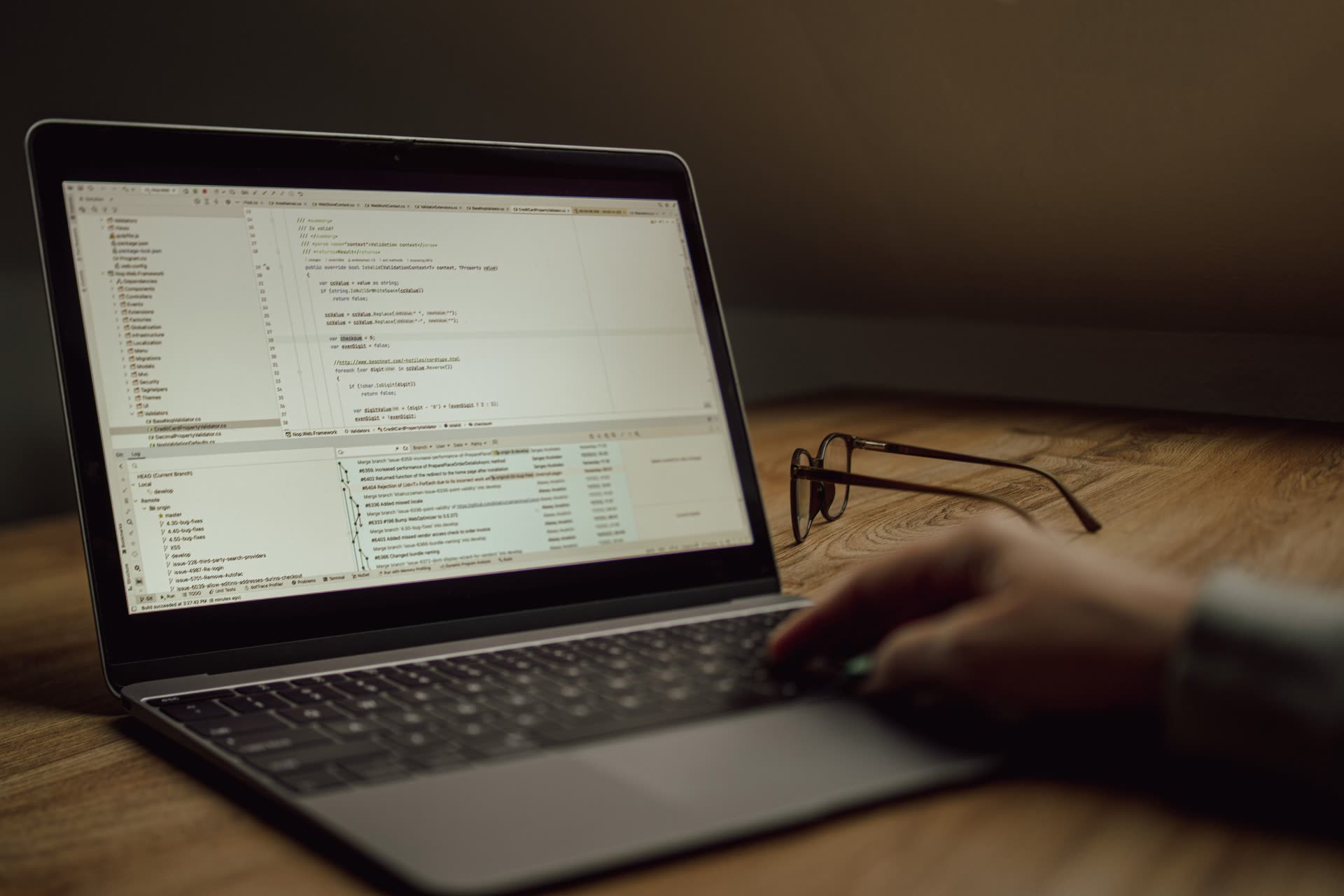 A hand on a laptop showing code, with reading glasses on a wooden desk in low light.